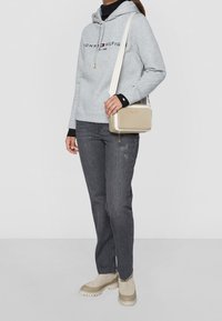 Sudadera gris claro con logo, camiseta de manga larga negra debajo, jeans oscuros, botas cortas marrones con suela gruesa y bolso bandolera beige con detalles dorados.