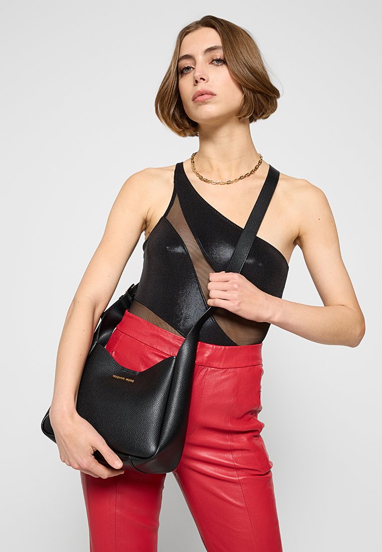 Schwarze Leder-Schultertasche mit gebogener Form und strukturiertem Finish. Modell trägt ein schwarzes Oberteil und rote Lederhosen. Einfaches Trägerdesign.