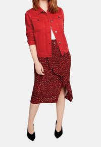 Rød denimjakke over en hvid top, parret med en knælang rød nederdel med et sort og guld blomstermønster og et sidelæg. Sorte hæle.