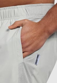 Pantalones cortos deportivos de color gris claro con una textura suave, que cuentan con un bolsillo lateral, cintura elástica y una etiqueta de logo negra en el dobladillo.