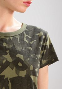 Olivově zelené tričko s kulatým výstřihem a maskáčovým vzorem, krátké rukávy a viditelné švy. Vyrobeno z bavlněné tkaniny.