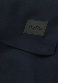 Tessuto a maglia blu scuro con colletto ripiegato, caratterizzato da un'etichetta rettangolare nera impresso con "HUGO". Texture liscia e design minimalista.