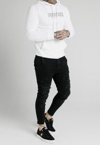 Vit huvtröja med grafisk text, kombinerad med svarta slitna jeans med färgstänk och svarta sneakers med vita sulor.