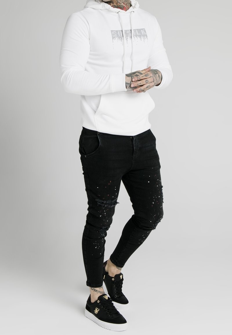 Vit huvtröja med grafisk text, kombinerad med svarta slitna jeans med färgstänk och svarta sneakers med vita sulor.