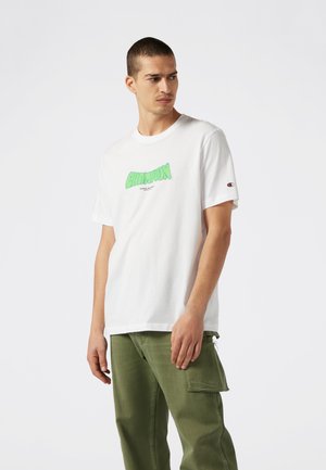 Maglietta bianca con una grafica verde "CHAMPION" sul davanti e un piccolo logo sulla manica; abbinata a pantaloni verdi.
