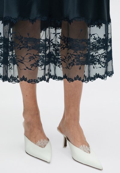 Zwarte satijnen jurk met een transparante kanten zoom met bloemmotieven. Witte mules met een puntige neus, kantaccenten en een matige hak.