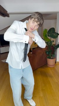 Cămașă albă cu mâneci lungi și cravată neagră, blugi largi de culoare bleu deschis și adidași albi; ambient interior cu o plantă verde și o canapea maro.