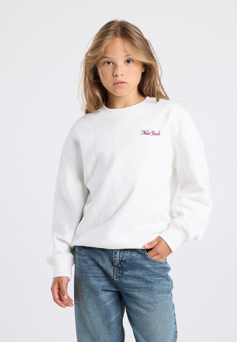 Jeune personne avec de longs cheveux blonds portant un sweat-shirt blanc avec le texte "New York" et un jean bleu, se tenant avec une main dans la poche.