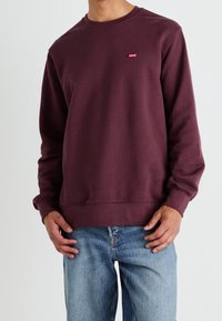Mörkröd sweatshirt med rund halsringning och ribbade ärmslut, med en liten röd logotyp på bröstet, i kombination med ljusblå jeans.