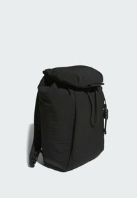 Sac à dos en tissu noir doux avec rabat supérieur, fermeture à lacet sur le devant et bretelles rembourrées, debout sur un fond clair.