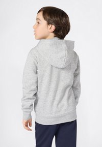 Ragazzo giovane che guarda a sinistra, indossa un hoodie grigio e pantaloni scuri su uno sfondo bianco semplice.