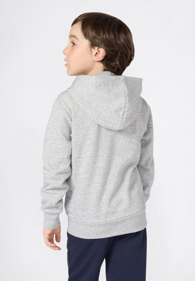 Ragazzo giovane che guarda a sinistra, indossa un hoodie grigio e pantaloni scuri su uno sfondo bianco semplice.
