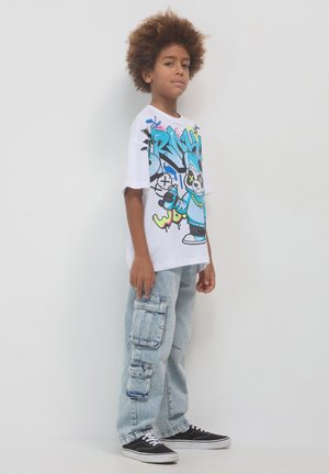 T-shirt grafica bianca con stampa colorata, pantaloni cargo in denim azzurro chiaro con molte tasche e sneaker nere con lacci bianchi.