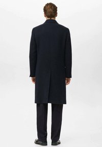 Lungo cappotto blu navy con taglio dritto, spacco posteriore singolo e colletto a intaglio. Realizzato in una miscela di lana texturizzata, indossato sopra pantaloni scuri e scarpe.