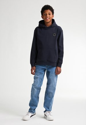 Felpa navy con tasca frontale e patch con logo, abbinata a jeans azzurri e sneakers bianche. Il tessuto sembra morbido e lavorato.