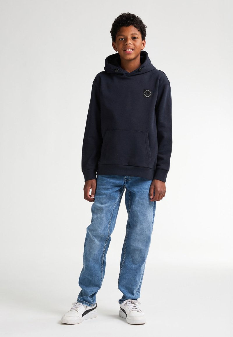 Felpa navy con tasca frontale e patch con logo, abbinata a jeans azzurri e sneakers bianche. Il tessuto sembra morbido e lavorato.