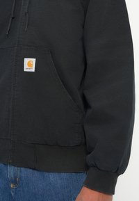 Sort Carhartt hoodie med frontlomme og snore, iført med blå denim jeans, der viser delvist torso og arm.
