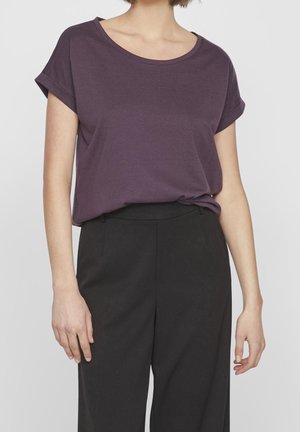 Osoba ubrana w fioletową koszulkę z krótkim rękawem wpuszczoną do czarnych spodnie z wysokim stanem, stojąca z rękami swobodnie opuszczonymi wzdłuż ciała.