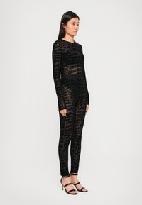 Juodas permatomas bodysuit su ilgomis rankovėmis, puošiamas viso paviršiaus Versace logotipo raštu. Minkšta tekstūra ir prigludęs dizainas, derinamas su kryžminiais aukštakulniais.