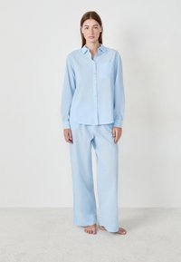Mujer de pie descalza sobre una alfombra clara, vestida con una camisa de botones azul claro y pantalones de pierna ancha a juego, contra un fondo blanco liso.