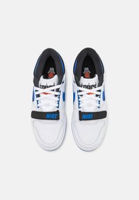 Zapatillas blancas con acentos azules, cuero perforado texturizado, forro negro, cordones blancos y una correa negra con "NIKE" en azul.