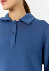 Zils polo krekls, kas izgatavots no mīksta auduma, ar apkakli, trim pogām un smalku logo izšuvumu uz krūtīm.