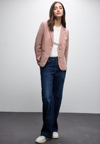 Růžový kordurový blazer se dvěma knoflíky, kombinovaný s bílým tričkem a tmavě modrými rozšířenými džíny. Bílá sportovní obuv celý outfit doplňuje.