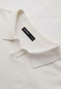 Witte polo met een geribbelde kraag en korte knoopsluiting, gemaakt van zacht stof, met een zwart label met "ombre" en maat "M."