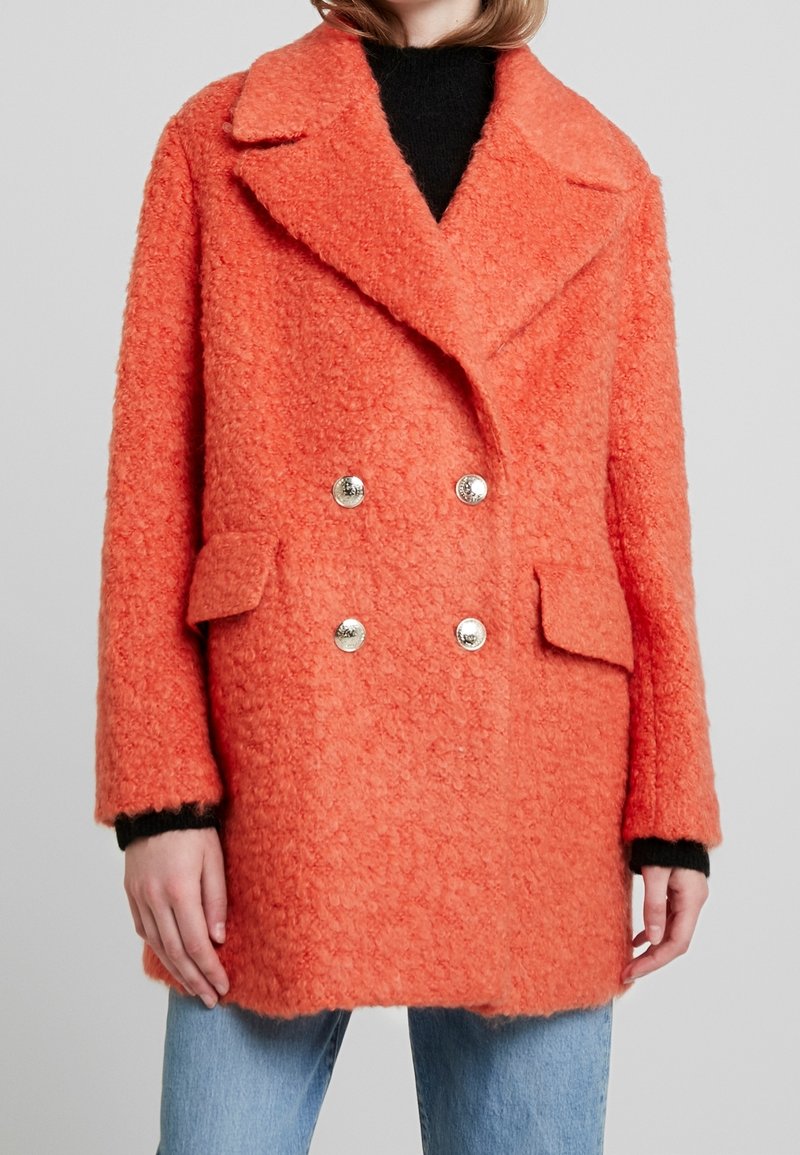 Manteau croisé en orange vif, fabriqué à partir d'un tissu texturé. Présente des boutons en métal argenté, un col à cran et deux poches avant.