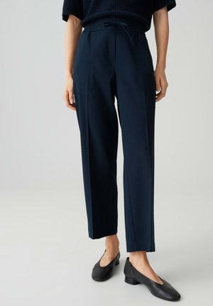 Personne portant un pantalon bleu marine sur mesure avec une taille à cordon et des chaussures noires à petit talon, debout devant un fond clair uni.