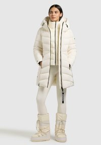 Witte pufferjas met een capuchon en een rits aan de voorkant, met een doorgestikte textuur. Ggedragen over bijpassende crèmekleurige leggings en snowschoenen met veters.