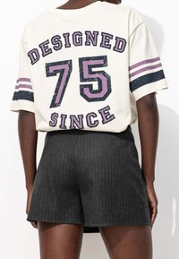 Vue arrière d'une personne portant une chemise crème avec le texte "Conçu 75 Depuis" et des manches rayées, associée à un short à fines rayures sombres.