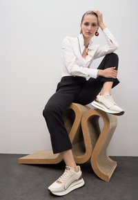 Camisa blanca de botones con detalles dorados, pantalones negros ajustados y zapatillas crema con detalles dorados, sentado en una silla con textura ondulada.