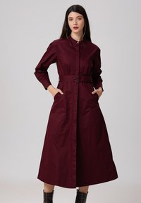 Robe longue bordeaux à manches longues avec une taille ceinturée, présentant un col boutonné et des poches latérales. Tissu lisse avec une silhouette droite.