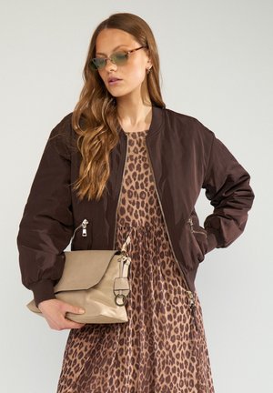 Frau mit langen Haaren, die Sonnenbrillen trägt, eine dunkelbraune Bomberjacke, ein Kleid mit Leopardenmuster und eine beige Handtasche vor einem schlichten Hintergrund hält.