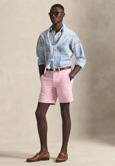 Rutig knappskjorta i ljusblå och gröna toner, rosa shorts, brunt skärp, bruna loafers och en ljusblå tröja hängande över axlarna.