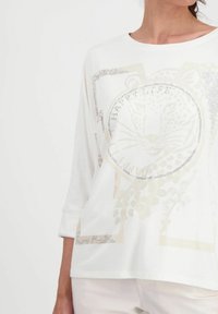 Frau trägt ein weißes Langarmshirt mit einem mit Strass verzierten Leopardenkopf und dem Text "HAPPY LIFE" auf der Vorderseite.