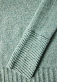 Ljusgrön tröja med mjuk textur, med ribbade manschetter och fåll. Tyget visar ett diskret, melerat mönster.