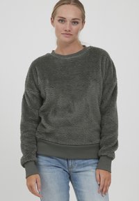 Felpa grigia in pile con una texture soffice, polsini e orlo a coste. Collo rotondo e vestibilità rilassata, abbinata a jeans azzurri chiari.