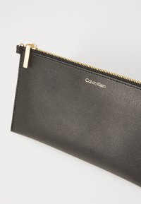 Svart lær clutch med gullglidelås og Calvin Klein-merke, plassert mot en lys bakgrunn.