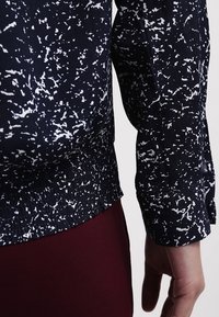 Gros plan sur le bras d'une personne portant un haut à manches longues noir et blanc à pois, avec un bas resserré, à côté d'un pantalon rouge foncé.