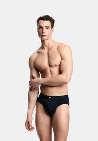 Męskie slipy w granatowym kolorze z gładkiej tkaniny, o dopasowanym kroju i paskiem w talii z logo. Model prezentuje się z neutralnym wyrazem twarzy.