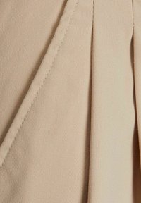 Béžová látka s jemnou texturou, obsahující záhyby a precizní šití. Materiál se jeví jako lehký a hladký.