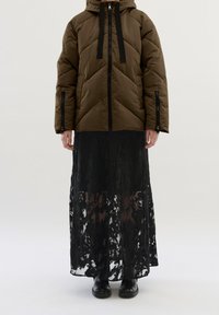 Brązowa puchowa kurtka z kapturem, z czarnymi sznurkami i zamkami błyskawicznymi; nałożona na długą czarną koronkową spódnicę. Czarne botki za kostkę dopełniają stylizację.