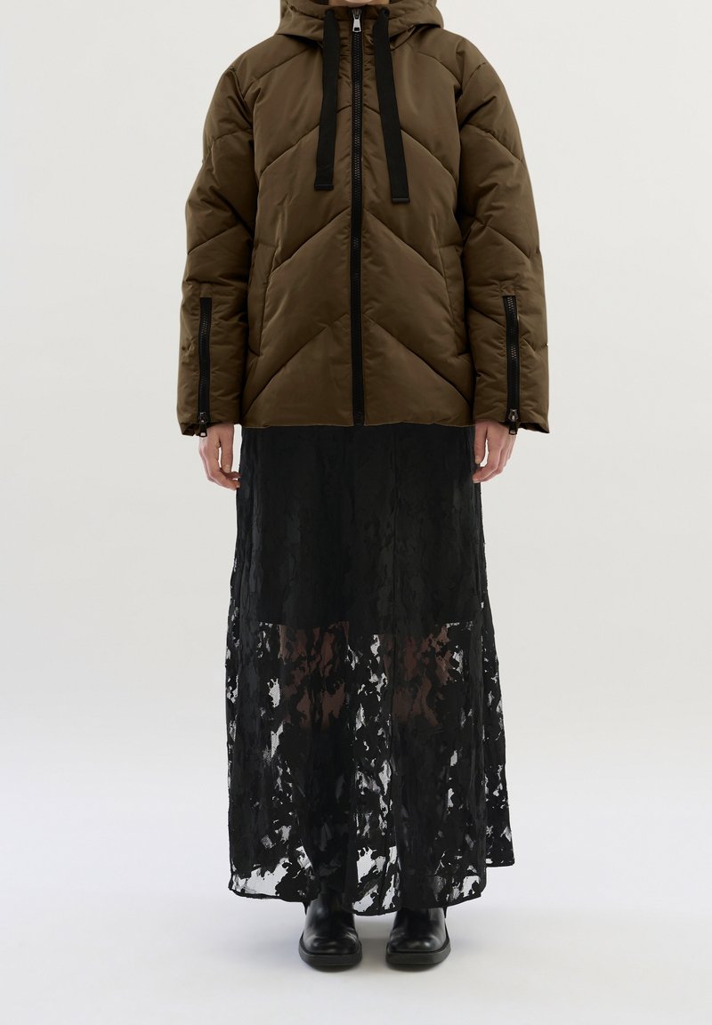 Brązowa puchowa kurtka z kapturem, z czarnymi sznurkami i zamkami błyskawicznymi; nałożona na długą czarną koronkową spódnicę. Czarne botki za kostkę dopełniają stylizację.