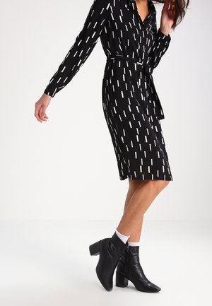 Kobieta ubrana w czarną sukienkę z białymi prostokątnymi wzorami, w białych skarpetkach i czarnych botkach na średnim obcasie, stojąca na białej podłodze.