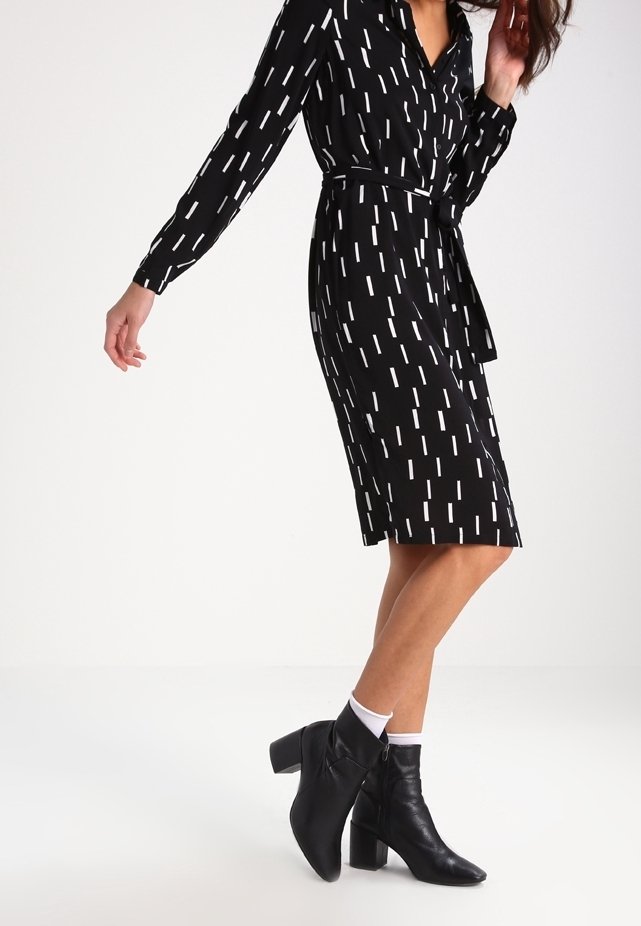 Femme portant une robe noire avec des motifs rectangulaires blancs, associée à des chaussettes blanches et des bottines noires à talon moyen, debout sur un sol blanc.