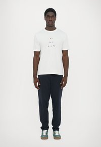 White cotton t-shirt featuring colorful text "PS PAUL SMITH" on the front, paired with navy pants and green sneakers.