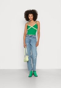 Top verde com detalhes em renda, jeans claros rasgados, botins verdes à altura do tornozelo e uma pequena bolsa verde. Fundo plano, tons neutros.