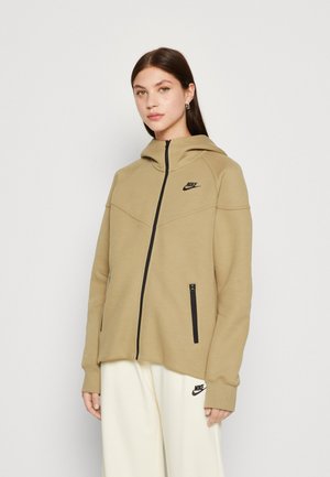 Felpa Nike beige con zip e cappuccio, logo nero e due tasche con zip nere. Texture liscia e vestibilità atletica.